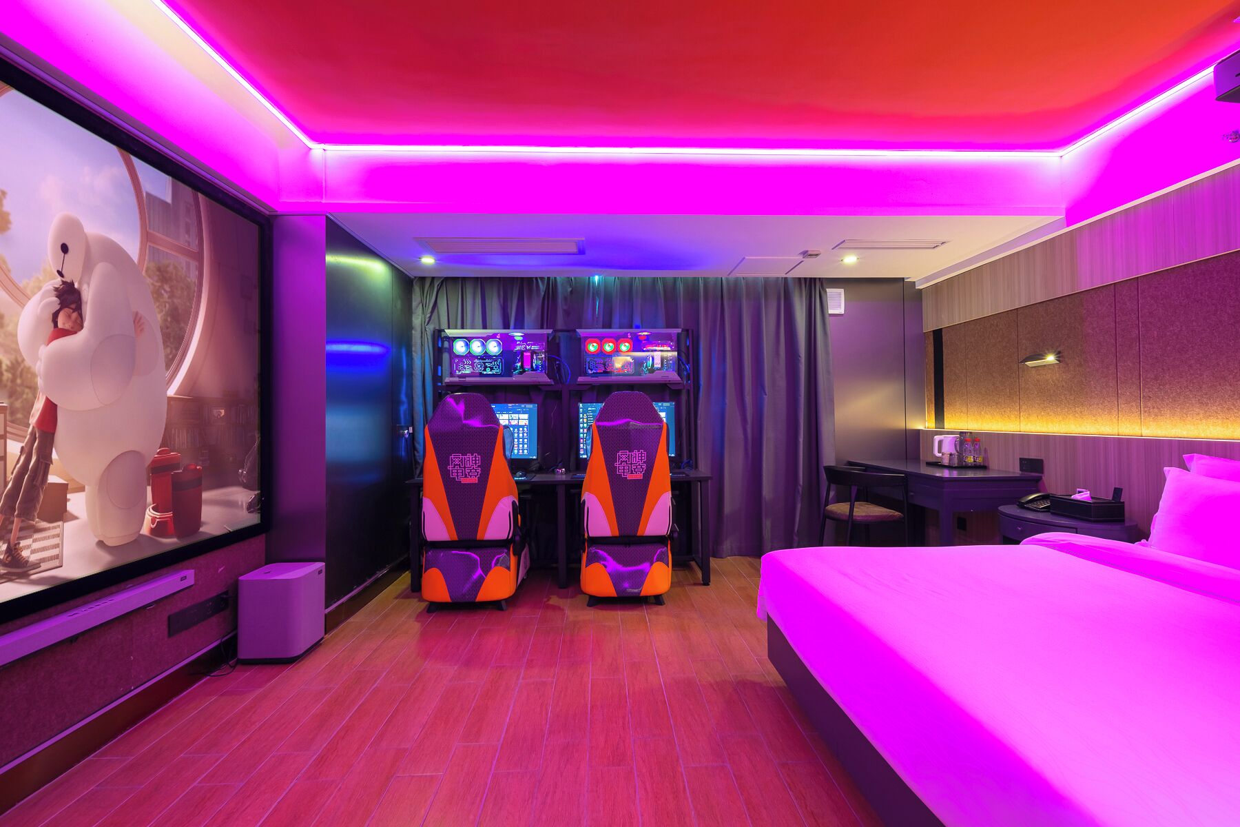 A modern hotel room with a large bed, a desk with a computer, two gaming chairs, and a large screen displaying a video game.