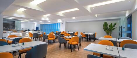 A modern dining area with multiple tables and chairs, a kitchen in the background, and a large plant.