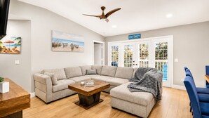 A living room with a sectional sofa, a coffee table, and a ceiling fan. There is a door with glass panels and a sign above it.