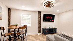 A modern living room with a dining area, a flat-screen TV mounted on the wall, and a wooden dining table with chairs.