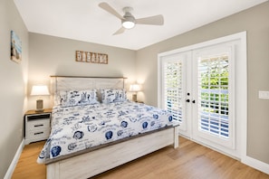 Quarto com cama, duas mesas de cabeceira, um ventilador de teto e uma porta com vista para o verde.