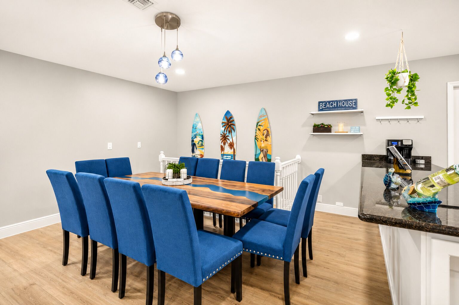 Ein Essbereich mit Holztisch und blau gepolsterten Stühlen, eine Küche mit schwarzer Arbeitsplatte und Surfbrettern an der Wand.