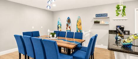 Ein Essbereich mit Holztisch und blau gepolsterten Stühlen, eine Küche mit schwarzer Arbeitsplatte und Surfbrettern an der Wand.