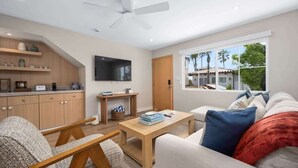 A modern living room with a sofa, coffee table, and a TV mounted on the wall. There is a wooden cabinet with shelves and a window providing a view outside. - Modern 2BR Near Beach w/ Parking, Spa, BBQ, EV (San Diego)