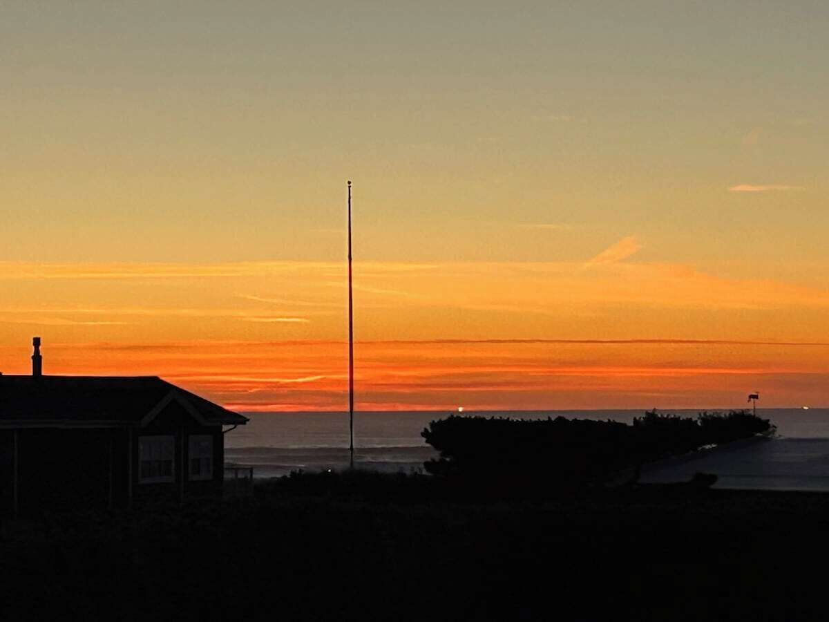 Un atardecer costero con la silueta de una casa, un asta de bandera y un muelle.