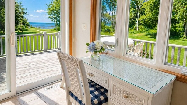 A room with a glass desk, a wicker chair, a view of the sea, and a white railing.