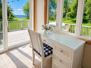 A room with a glass desk, a wicker chair, a view of the sea, and a white railing.