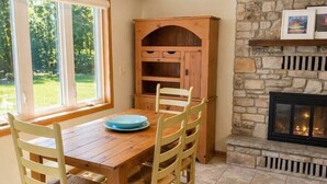 A dining area with a wooden table and chairs, a stone fireplace, and a window with a view of greenery.