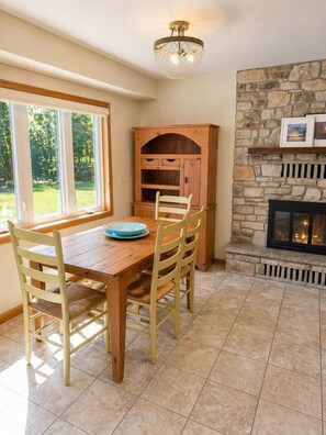 A dining area with a wooden table and chairs, a stone fireplace, and a window with a view of greenery. - Waterfront Home|Dock & Kayaks|5 Min to Baileys Harbor (Baileys Harbor)