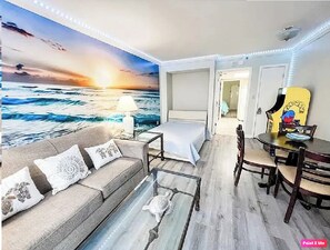 A room with a bed, a sofa, a dining table, and a mural of a sunset over the ocean. - Oceanfront Paradise with Private Balcony! (Myrtle Beach)