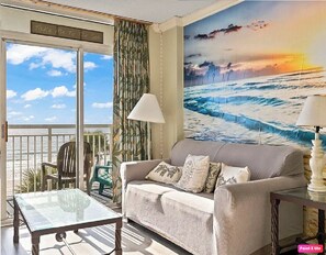 A living room with a large window overlooking the sea, a sofa with cushions, a glass table, and a painting of ocean waves. - Oceanfront Paradise with Private Balcony! (Myrtle Beach)