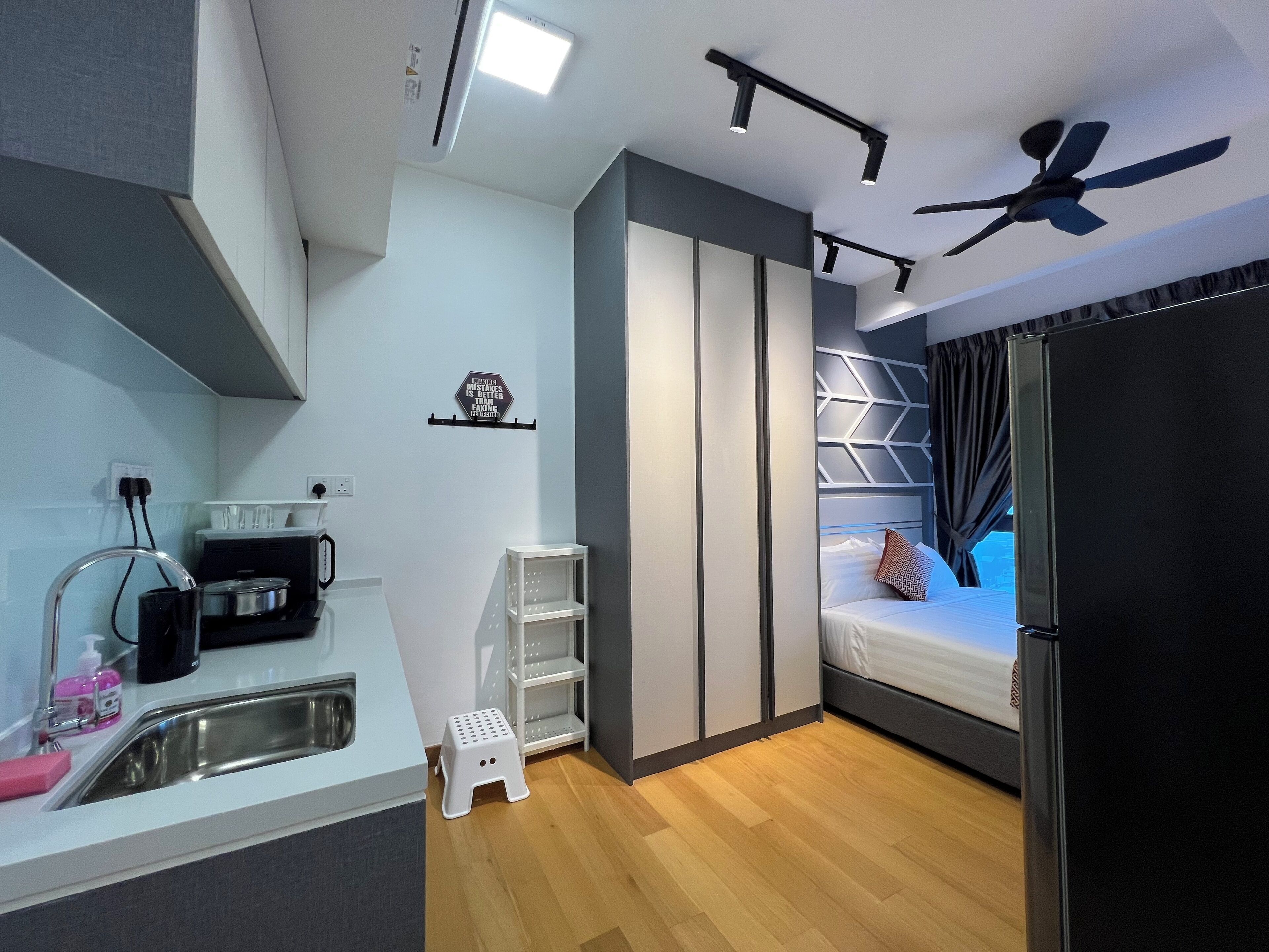 A compact hotel room with a kitchenette, a bed with a headboard, a ceiling fan, and a small stool.