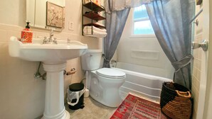 A bathroom with a pedestal sink, toilet, and bathtub. There is a shower curtain, a small shelf with toiletries, and a red patterned rug. - Sweet Home with Ample Parking. Only 1-min to Freeway! (Sacramento)