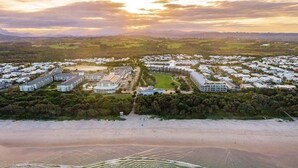 An aerial view of a coastal resort with multiple buildings, a beachfront, and a hilly landscape in the background. - Palm Horizon -3B Bali Resort in Kingscliff (1218) (Tweed)