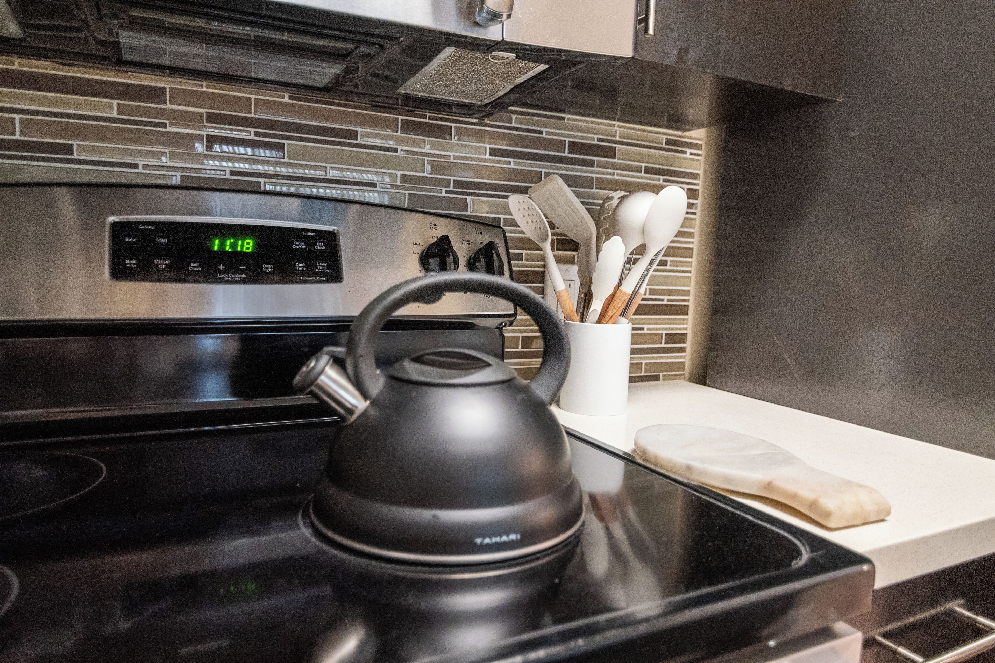 A kitchen with a stainless steel oven, a black electric kettle, and a white utensil holder on the counter.
