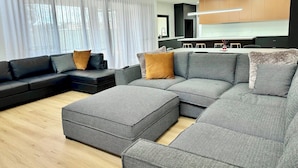 A modern living room with a sectional sofa, ottoman, and a dining area in the background. - Emmylou Escape - Sleeps 16 (Moama)