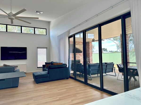 A modern living room with a flat-screen TV, a ceiling fan, and a large sliding glass door leading to an outdoor patio with a brick fireplace.