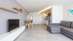 A modern living room with a flat-screen TV, a grey sofa, and a dining area with a table and chairs. - Ocean View Apt - 1BR T2-613 (Chalong)