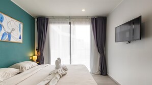 A hotel room with a bed, a painting on the wall, a TV mounted on the wall, and a window with curtains. - Ocean View Apt - 1BR T2-613 (Chalong)