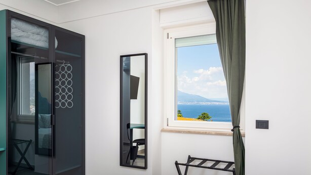 A bedroom with a bed, a desk, a chair, a wardrobe, a mirror, and a window with a view of the sea and mountains. - Maison Regina Giovanna (Sorrento)
