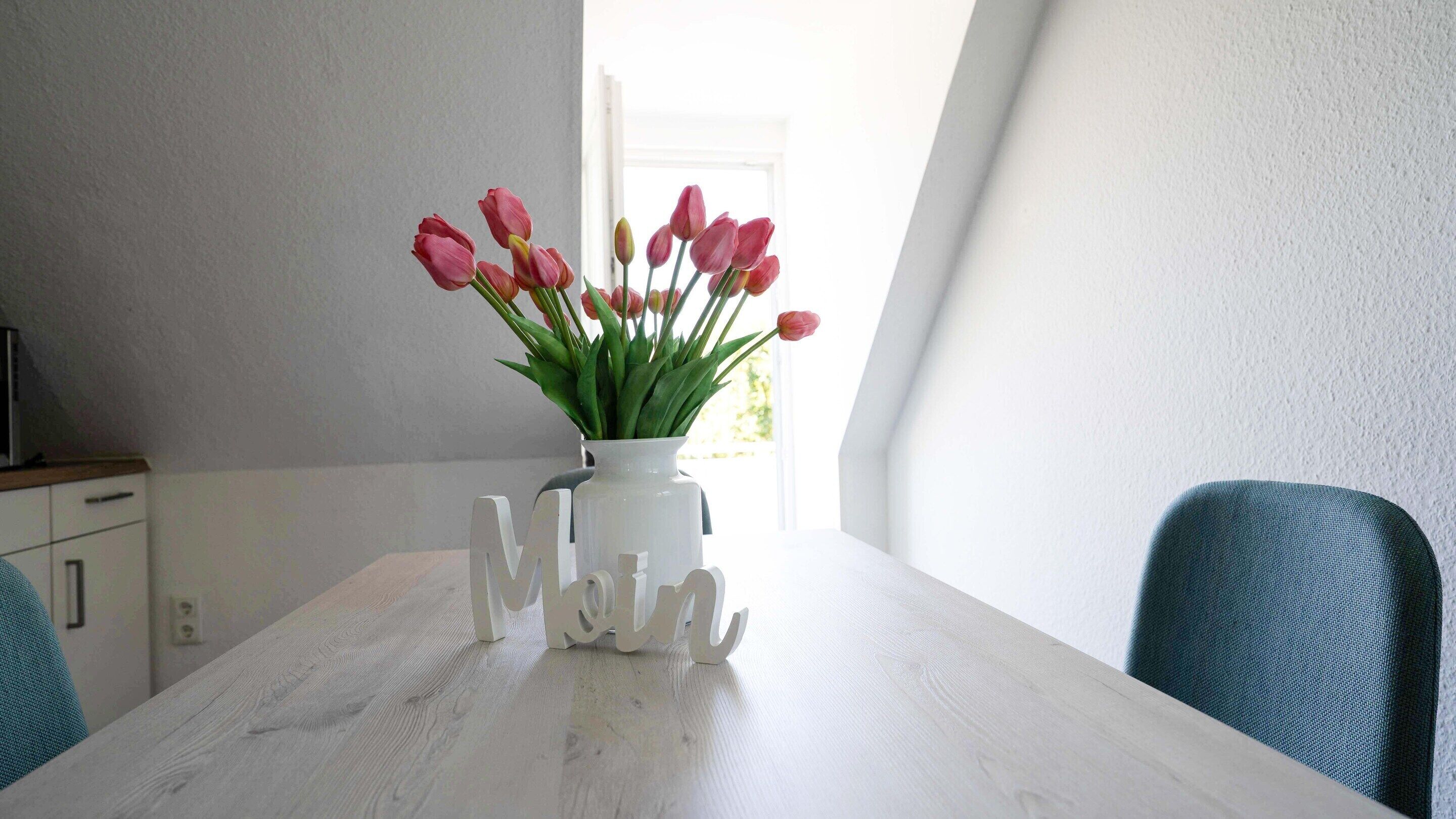 Una mesa de comedor con un jarrón blanco con tulipanes rosados, un jarrón blanco con la palabra “MOM” en cursiva y sillas color turquesa.