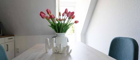 Una mesa de comedor con un jarrón blanco con tulipanes rosados, un jarrón blanco con la palabra “MOM” en cursiva y sillas color turquesa.