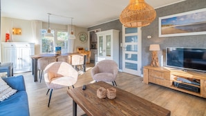 A spacious living room with a wooden coffee table, two armchairs, a sofa, a TV, and a dining area with a table and chairs. - Charming Villa Near the sea and Forest (De Koog)
