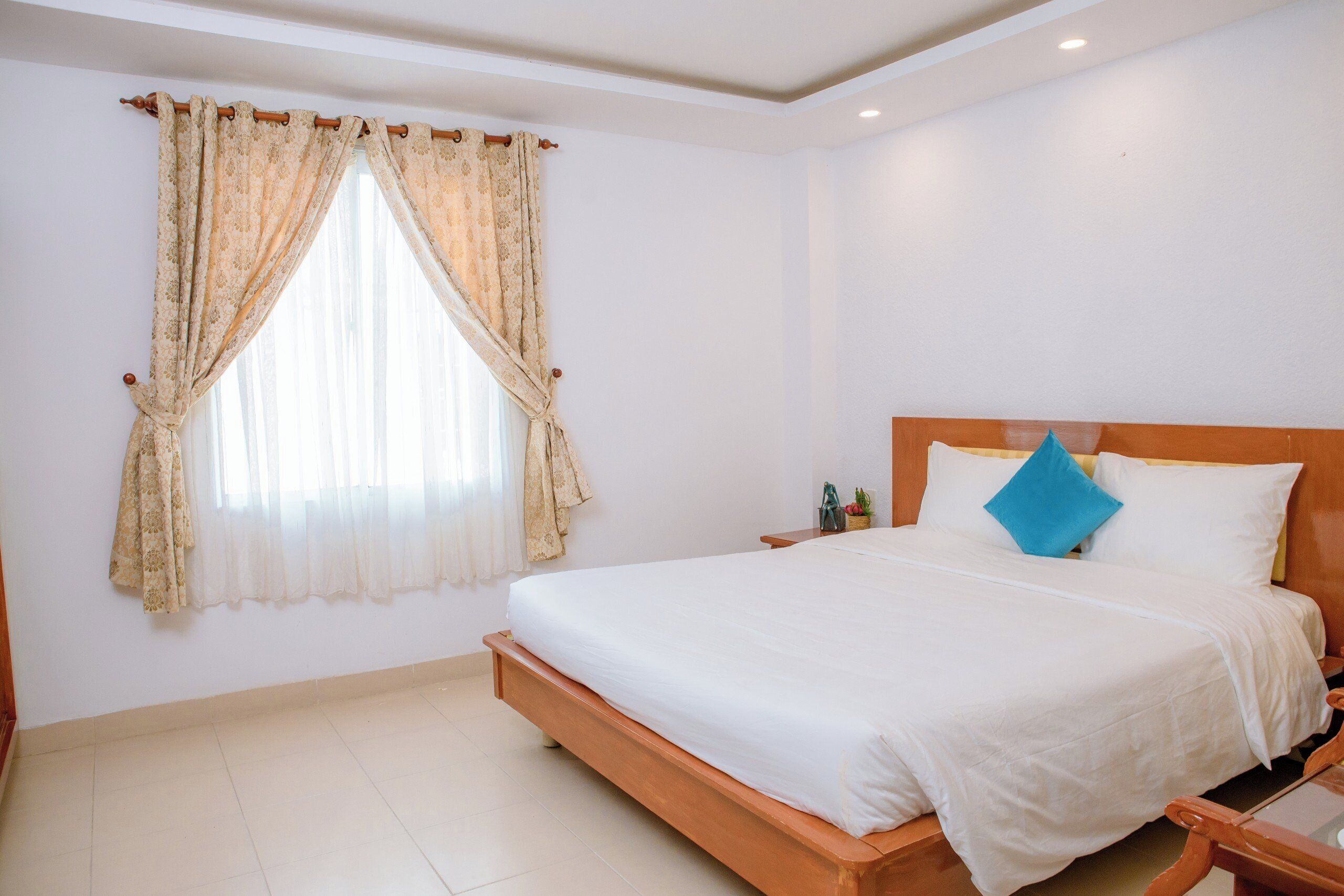 A bedroom with a wooden bed, white bedding, a window with sheer curtains, and a nightstand with a lamp.