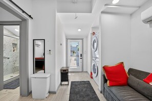 A laundry room with a washing machine, a trash bin, a mirror, and a grey sofa with a red pillow. - FIFA World Cup 2026 Retreat | Leimert Park (Los Angeles)