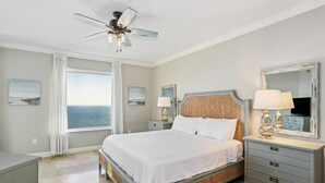 A bedroom with a bed, nightstands, a dresser, a ceiling fan, and a large window with a view.