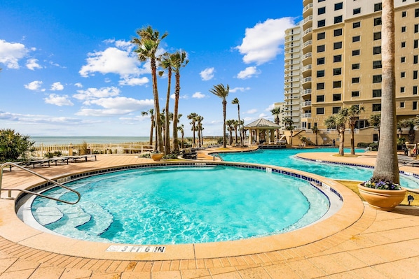 A pool area with multiple circular pools, palm trees, and a high-rise building in the background.