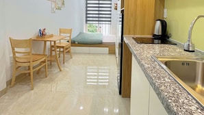 A modern kitchen with a granite countertop, stainless steel sink, and wooden cabinets. There is a dining area with a table and chairs, and a window with blinds. - Hat Nang House - Go Vap (Ho Chi Minh City)