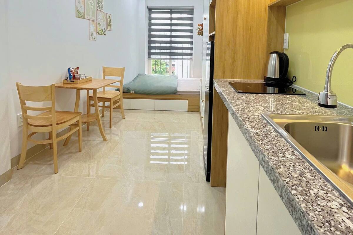 A modern kitchen with a granite countertop, stainless steel sink, and wooden cabinets. There is a dining area with a table and chairs, and a window with blinds.