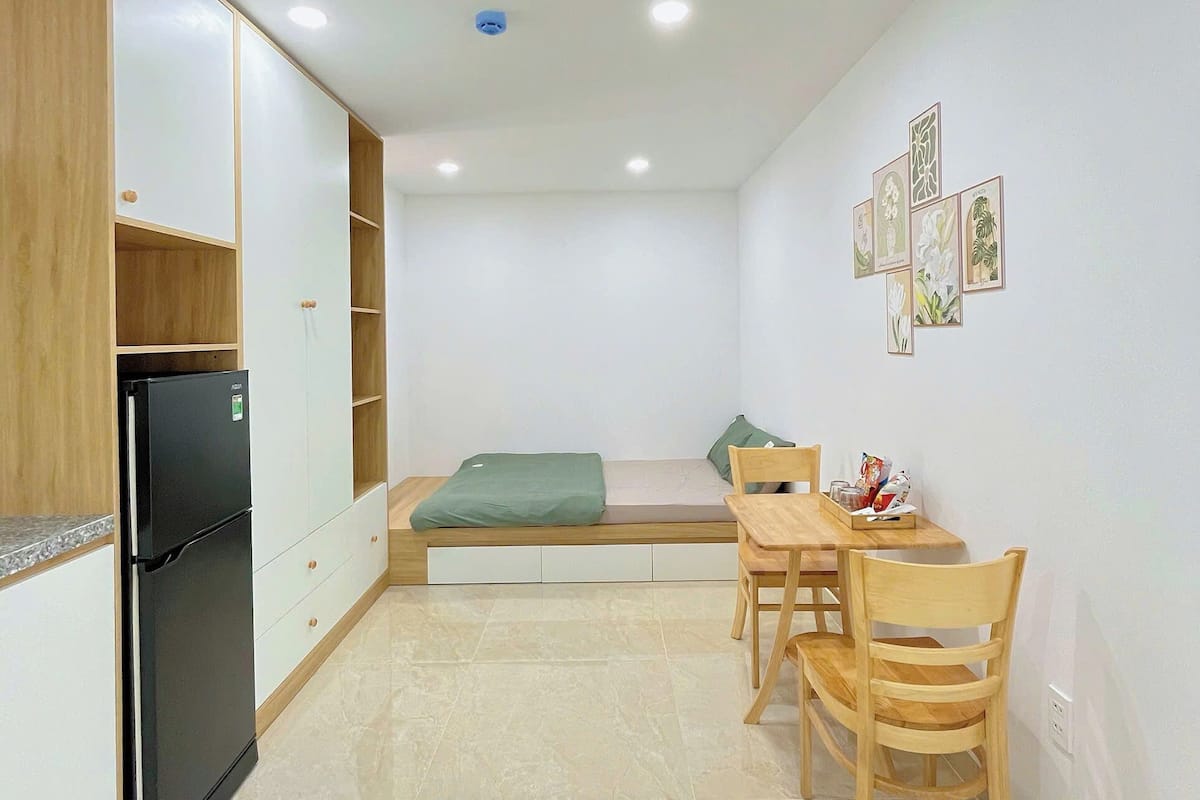 A compact kitchen with a refrigerator, wooden cabinets, a dining table with chairs, and a small bed.