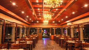 Un restaurante con vigas de madera en el techo, candelabros y ventanas amplias. Las mesas están listas con platos y copas.