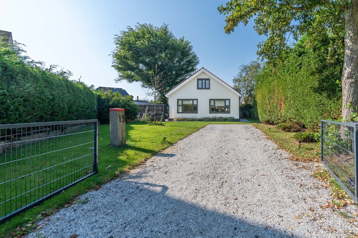 A house with a gravel driveway leading to a white exterior, surrounded by greenery and a metal fence.