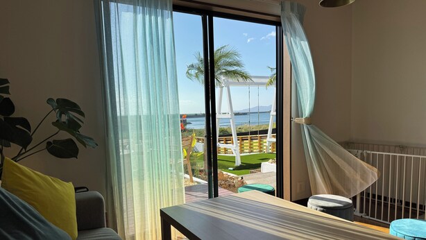 A room with a sofa, a table, and a view of a garden and water body through large windows. - Aoniwa - AWAJI (Awaji)