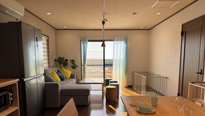 A modern kitchen with a built-in refrigerator, wooden dining table, and a seating area with a sofa and ottoman. - Aoniwa - AWAJI (Awaji)