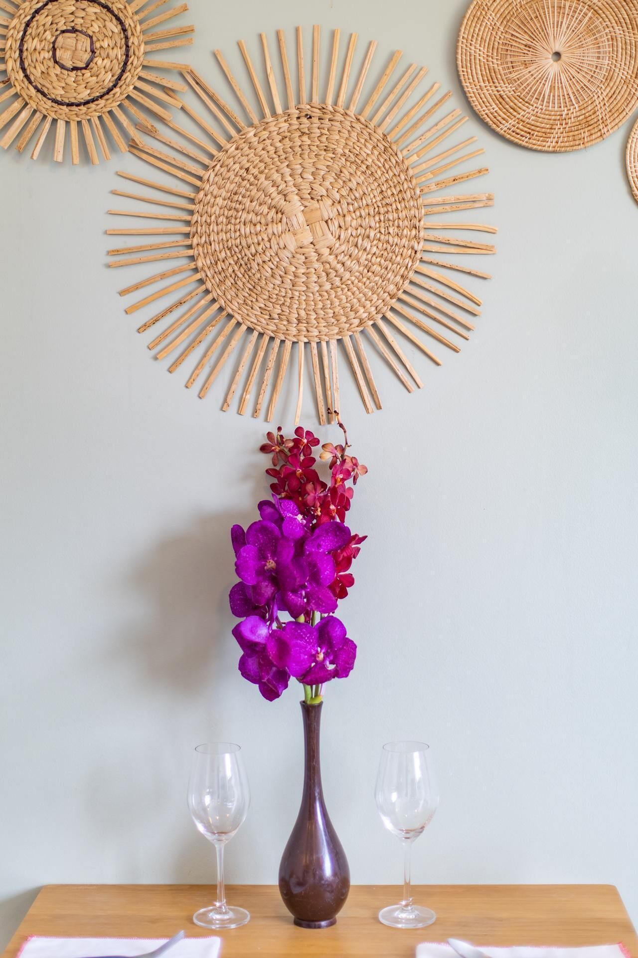 Una mesa de comedor con un jarrón de flores moradas, dos copas de vino y un adorno de pared tejido.