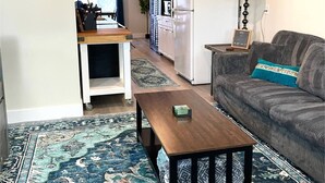 Ein Wohnzimmer mit einer Couch, einem Couchtisch, einem Teppich und einer Küchenzeile mit einem Kühlschrank sowie einer Uhr an der Wand.