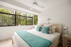 Une chambre avec une grande fen être, un lit recouvert d’une couverture turquoise, une table de chevet et un ventilateur de plafond.