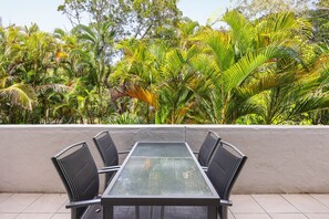 Un espace de restauration en plein air avec une table à plateau de verre et des chaises noires, entouré d’un mur blanc et d’une végétation tropicale.