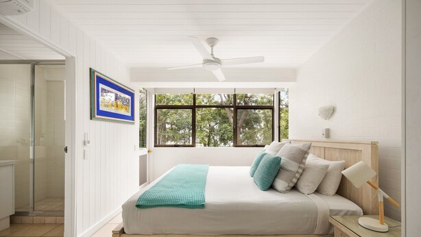 A bedroom with a bed, a nightstand, a window with a view of trees, and a ceiling fan. - Tea Trees 5 - Coastal Calm near Hastings St (Noosa Heads)