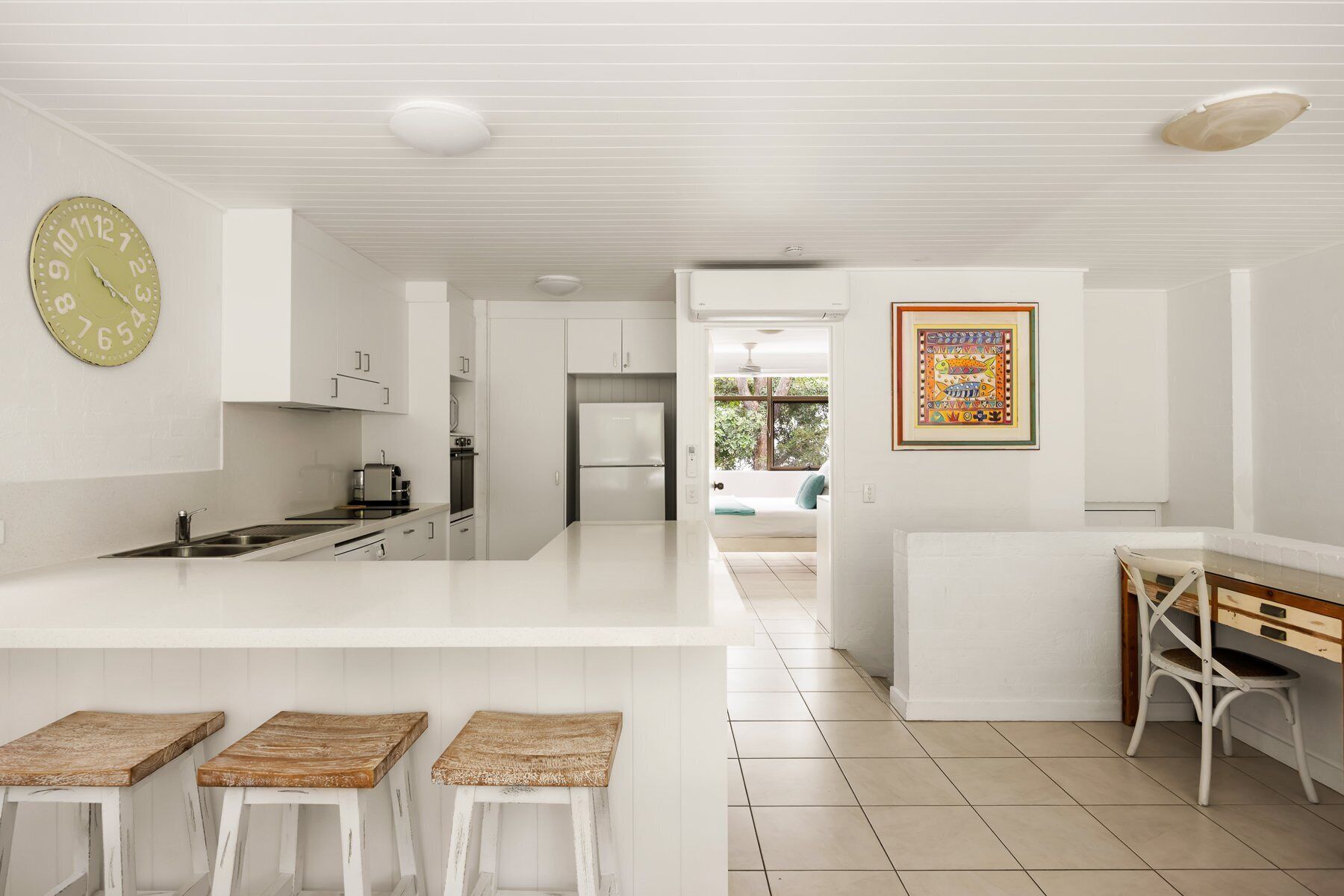Una cocina moderna con muebles blancos, una isla central con taburetes, un reloj y un cuadro en la pared.