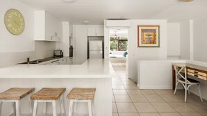 A modern kitchen with white cabinetry, a central island with stools, a clock, and a framed artwork on the wall. - Tea Trees 5 - Coastal Calm near Hastings St (Noosa Heads)