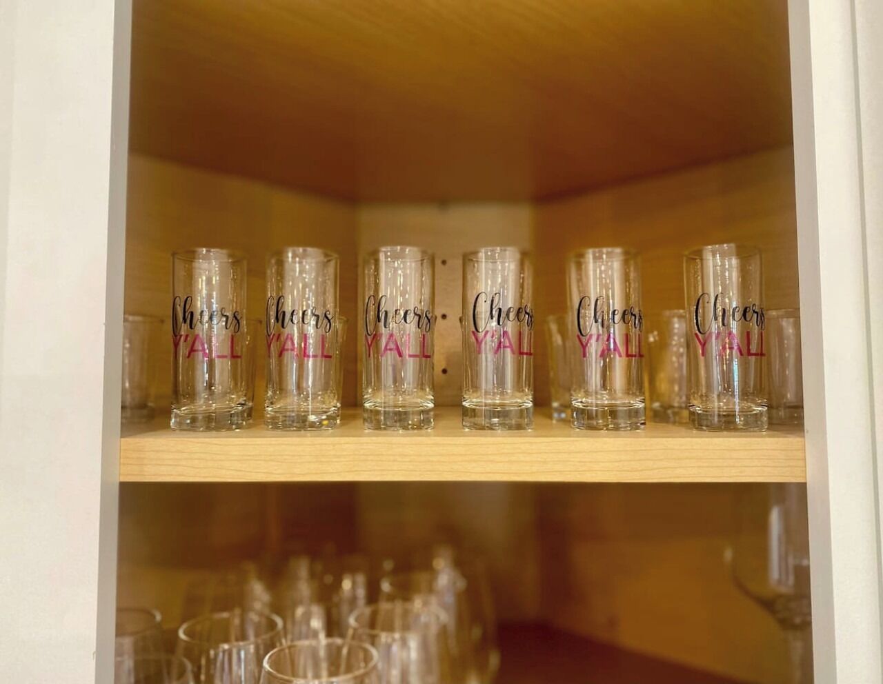 Una estantería con varios vasos transparentes que dicen "Cheers Y'ALL" en una vitrina de madera.