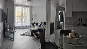 A modern living room with a glass coffee table, a sofa, and a dining area with a glass table. There is a kitchen area with white cabinets and a sink.
