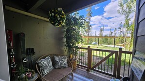 A covered porch with a wicker sofa, a hanging flower basket, and a view of a forested area.