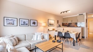 A modern living room with a sofa, a coffee table, and a dining area with chairs and a table. There are framed pictures on the wall and a kitchen in the background.