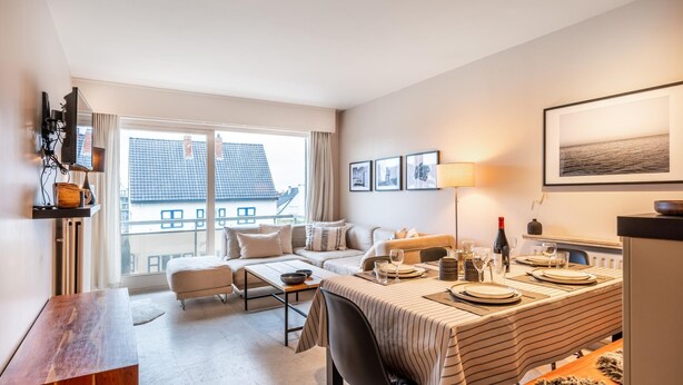 A modern hotel room with a dining area, a sofa set, and a large window offering a view of the cityscape.